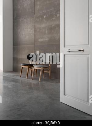 Zwei Stühle im Loft-Innenraum - Blick von einem Flur. Ein paar Holzstühle auf Betonboden in der Nähe einer leeren graubraunen Wand, innen mit hohen Decken Stockfoto