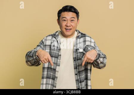 Abonnieren. Asiatischer Mann mittleren Alters, der nach unten zeigt und sagt, er solle sich Werbung ansehen, gestickt in Ordnung, zeigt unten einen Ort für kommerzielle Werbeinformationen. Reifer Chinese auf beigefarbenem Hintergrund Stockfoto