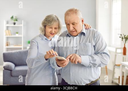 Seniorenpaar, das gemeinsam ein Mobiltelefon benutzt und zu Hause im Wohnzimmer steht. Stockfoto