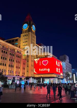Lebhafte Nacht in Peking mit dem berühmten Uhrenturm und der leuchtenden „i ❤️ Beijing“-Werbetafel in einer belebten Stadtstraße. Stockfoto