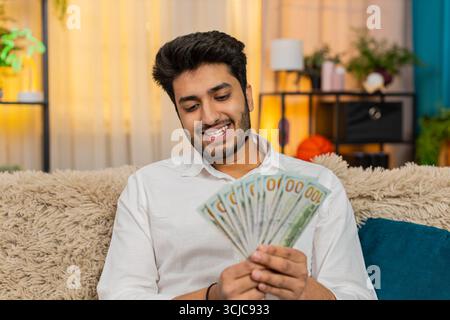 Indischer Mann zu Hause, der Bargeld zählt, sein Budget sorgfältig überlegt und zukünftige Ausgaben plant. Ein Araber sitzt auf dem Sofa und konzentriert sich auf Geldmanagement und durchdachte Finanzentscheidungen Stockfoto