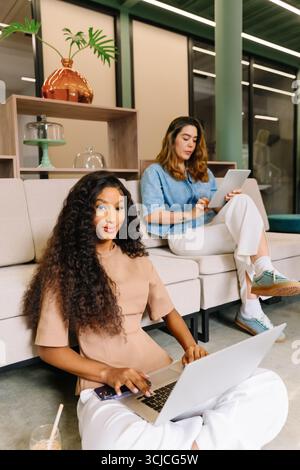 Zwei Frauen arbeiten in einem modernen Coworking-Raum zusammen und setzen sich mit Technologie auseinander. Der eine benutzt einen Laptop auf dem Boden, der andere sitzt mit einem Tablet, um zu fördern Stockfoto