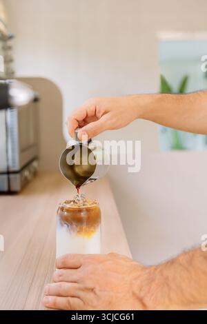 Eine Person gießt gekonnt Espresso über Eis und Milch in ein Glas und bastelt in einer hellen, modernen Küche einen erfrischenden Eiskaffee. Die Umgebung ist gemütlich und Stockfoto