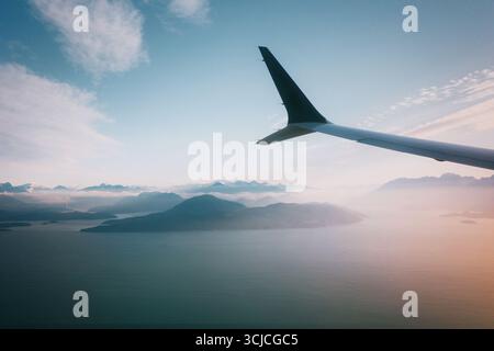 Eine malerische Aussicht aus einem Flugzeugfenster, mit dem Flügel vor dem Hintergrund majestätischer Berge und ruhiger Gewässer, unter einem klaren Himmel mit weichen Wolken Stockfoto