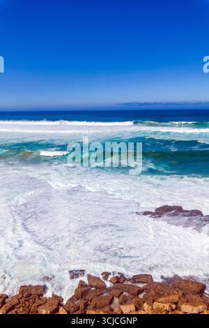 Mächtige Wellen krachen unter einem leuchtend blauen Himmel gegen zerklüftete Küstenfelsen, mit einem entfernten Gebäude an der Klippe mit Blick auf den Ozean Stockfoto