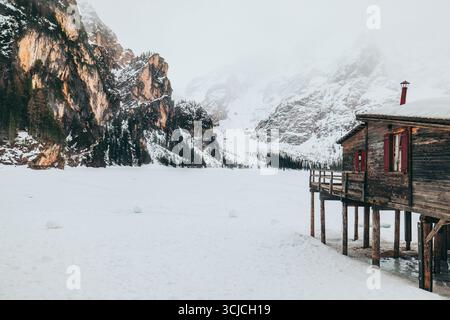 Eine atemberaubende Winterszene in den Dolomiten, Italien, mit einer rustikalen Hütte neben einem gefrorenen See, umgeben von schneebedeckten Bergen und Kiefern u Stockfoto