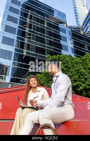 Zwei multikulturelle Fachleute führen in Melbourne eine lebhafte Geschäftsdiskussion im Freien. Sie sitzen auf einer roten Bank, mit einem Tablett, mit Wolkenkratzer Stockfoto