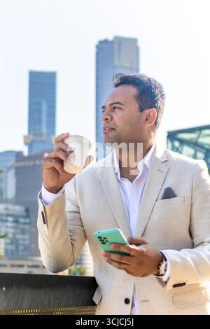 Ein professioneller Mann in einem leichten Anzug hält eine Kaffeetasse und ein Smartphone und steht draußen mit der Skyline von Melbourne im Hintergrund und reflektiert ein Ur Stockfoto