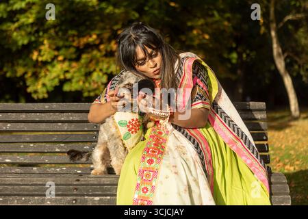 Indische Frau in traditioneller Kleidung, die auf einer Parkbank sitzt und ihren kleinen Hund umschließt, während sie einen sonnigen Herbsttag genießt. Die lebendigen Blätter führen zu einem Krieg Stockfoto