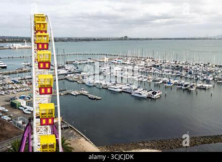 Drohnenblick auf die Uferpromenade von Geelong, mit einem pulsierenden Riesenrad und einem geschäftigen Yachthafen voller Boote, unter einem bewölkten Himmel, der die Stadt erfasst Stockfoto