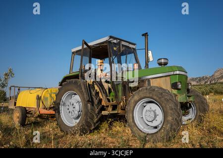 Ein älterer Mann mit Hut fährt einen Traktor mit Wassertank über eine sonnenbeschienene Farm. Das Bild zeigt einen traditionellen Lebensstil auf dem Land und ein modernes A Stockfoto