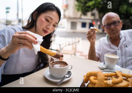 Eine Familie genießt spanische Churros, getaucht in heiße Schokolade in einem Café im Freien. Die Szene zeigt Wärme und Tradition vor einem pulsierenden Hintergrund der Stadt Stockfoto