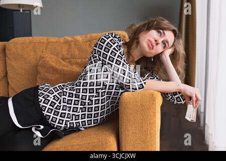 Eine junge Hausfrau entspannt sich auf einer gemütlichen Couch und blickt aus dem Fenster. Ihr ruhiger Ausdruck spiegelt einen friedlichen Moment in einem gemütlichen Zuhause wider Stockfoto