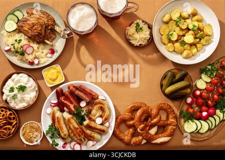 Blick von oben auf ein fest mit Schweinehaxe auf Sauerkraut, Bratwurst und Würstchen auf einem Teller, Brezeln, Kartoffelsalat, gegrillten Kartoffeln, Gurken, frisches Gemüse Stockfoto