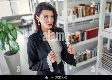 Eine selbstbewusste Frau in einem eleganten schwarzen Anzug posiert in einer Bibliothek und zeigt Eleganz und Professionalität. Die moderne Atmosphäre verstärkt sie Stockfoto