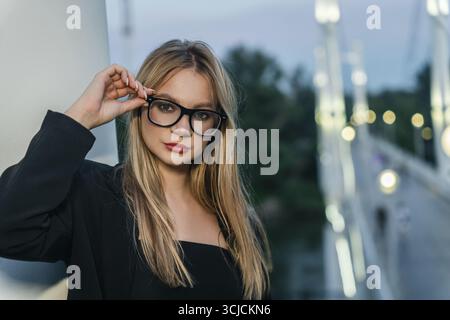 Junge blonde Frau mit schwarzer Brille, die draußen mit einem stilvollen schwarzen Outfit steht. Sie wirkt selbstbewusst und entspannt, während sie in den s posiert Stockfoto