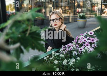 Junge blonde Frau mit Brille posiert im Freien, umgeben von grünem Laub und leuchtenden Blumen. Sie trägt eine schwarze Jacke, die moderne Raffinesse ausstrahlt Stockfoto