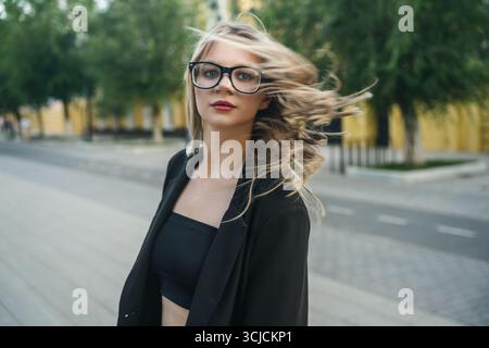 Die junge blonde Frau mit großer schwarzer Brille genießt einen luftigen Tag im Freien, ihr Haar fließt im Wind vor einem städtischen Hintergrund. Sie strahlt Vertrauen aus Stockfoto