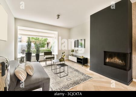 Ein stilvolles Wohnzimmer mit elegantem schwarzen Kamin, modernen Möbeln und gemustertem Teppich. Tageslicht füllt den Raum durch ein großes wi-fi Stockfoto