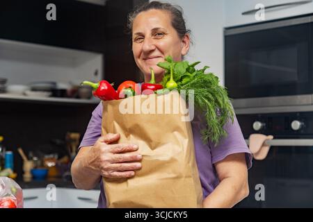Eine Frau mittleren Alters steht in ihrer gemütlichen Küche mit einem Papierbeutel gefüllt mit frischem Gemüse, einschließlich Paprika und Blattgemüse, und lächelt warm Stockfoto