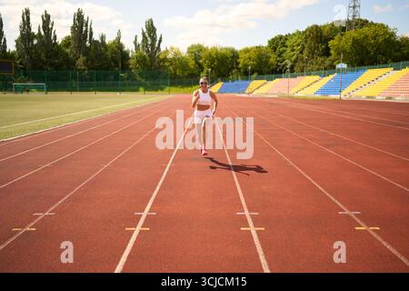 Eine Läuferin in sportlicher Ausrüstung joggt an einem sonnigen Tag auf einer Outdoor-Strecke. Ihr fokussierter Ausdruck und ihre dynamische Haltung verkörpern einen aktiven Lebensstil Stockfoto