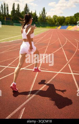 Eine fokussierte Läuferin in sportlicher Ausrüstung sprintet auf einer sonnendurchfluteten Outdoor-Strecke. Der helle Tag verstärkt ihre Energie und Entschlossenheit, während sie sich vorwärts bewegt Stockfoto