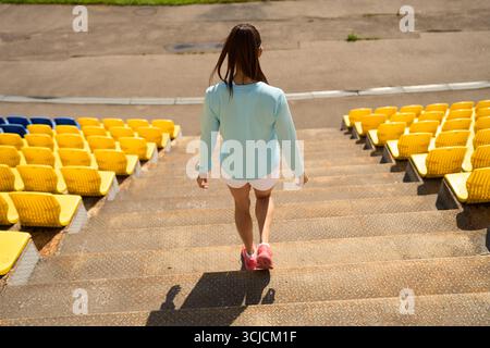 Eine Frau in Sportbekleidung geht selbstbewusst die Stadiontreppen hinauf und nimmt einen aktiven Lebensstil an. Die lebhaften gelben und blauen Sitzplätze sorgen für eine lebhafte Atmosphäre Stockfoto