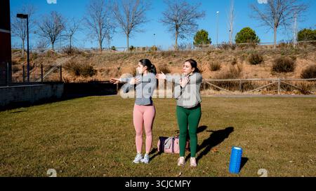 Zwei Frauen machen sich im Freien warm-up-Übungen und dehnen ihre Arme unter der Sonne. Sie stehen im Park, in sportlicher Kleidung, Stockfoto