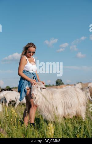 Eine lächelnde Frau mit Sonnenbrille und lässigem Outfit genießt einen sonnigen Tag auf einem Bauernhof, umgeben von Ziegen. Der klare blaue Himmel Stockfoto
