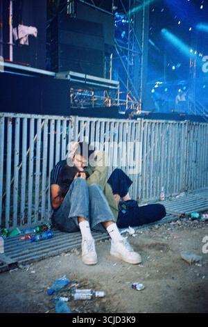 Junges Paar küsst sich neben der Hauptbühnenabsperrung des Festivals, sitzt auf dem Boden umgeben von Müll, aufgenommen mit direktem Analogblitz. Stockfoto