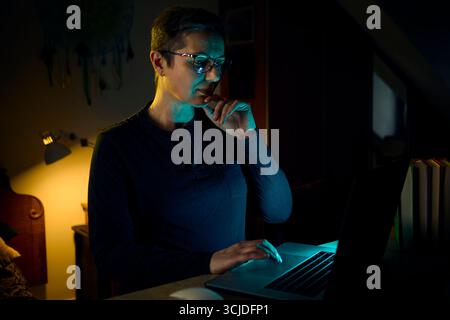 Eine Frau mittleren Alters arbeitet ferngesteuert an ihrem Laptop, während sie ihr Telefon hält und in einem dunklen Heimbüro multitaskt. Das Bild repräsentiert moderne Arbeiten aus HO Stockfoto