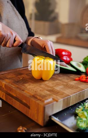 Eine Frau schneidet gelbe Paprika auf Holzbrett mit frischem Gemüse herum Stockfoto