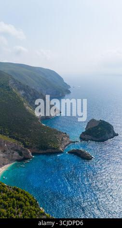 Blick aus der Vogelperspektive auf Agios Andreas Höhleninsel mit steilen felsigen Klippen, türkisfarbener Bucht, tiefblauem Ionischen Meer und grünen Hügeln, die sich bis zum Horizont erstrecken Stockfoto