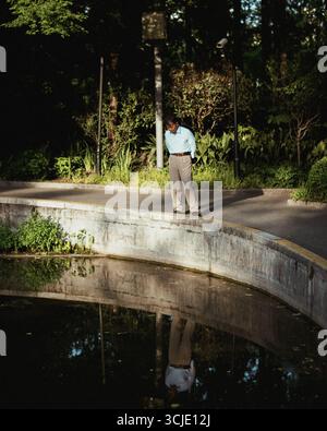 Älterer Mann, der in der Nähe eines Teichs im Shanghai Park steht, reflektiert im Wasser, besinnliche Stimmung Stockfoto