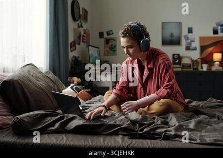 Junge Erwachsene kaukasische Frau mit einer Armprothese, die mit einem Laptop im Kreuz auf dem Bett sitzt und Kopfhörer trägt, wobei sie sich intensiv auf den Bildschirm im Schlafzimmer konzentriert und von persönlichen Gegenständen umgeben ist Stockfoto