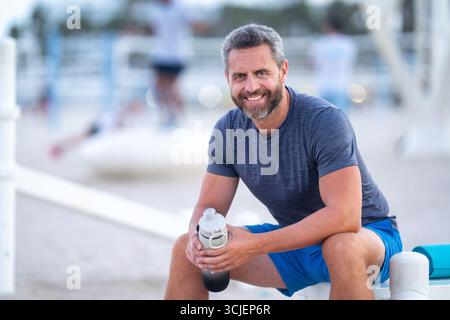 Sport-Fitness-Mann, der draußen auf einer Bank ruht. Reifes Fitnesssportmodell, das nach dem Training auf einer Bank sitzt. Hispanischer Sportmann macht danach eine Pause Stockfoto