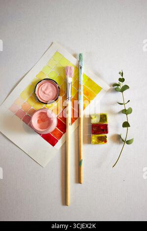 Kreative flache Lage aus Aquarellfarben, Pinseln und grünem Ast auf hellem Hintergrund. Künstlerischer Arbeitsbereich mit Pastellfarben und lebhaften Farben, perfekt für DE Stockfoto