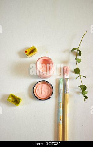 Kreative flache Lage aus Aquarellfarben, Pinseln und grünem Ast auf hellem Hintergrund. Künstlerischer Arbeitsbereich mit Pastellfarben und lebhaften Farben, perfekt für DE Stockfoto