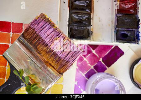 Kreatives Flachbild mit Aquarellpalette, Pinseln und Farbbehälter auf Papier. Künstlerischer Arbeitsbereich mit leuchtenden Farbfeldern in violetten, gelben und roten Tönen Stockfoto