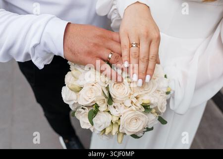 Hochzeitsringe an Händen mit Blumenstrauß, Hochzeitssymbolik, weiße Rosen, elegante Zeremonie, Liebe, Engagement, Romantik, Neutrale Töne, Nahaufnahme Stockfoto