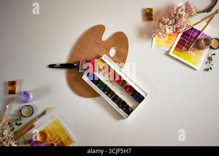 Künstlerische flache Lay mit Aquarellpalette, Pinseln, Farbmustern, Blumen und einer Holzpalette auf hellem Hintergrund. Kreativer Arbeitsbereich mit brillanter Leistung Stockfoto