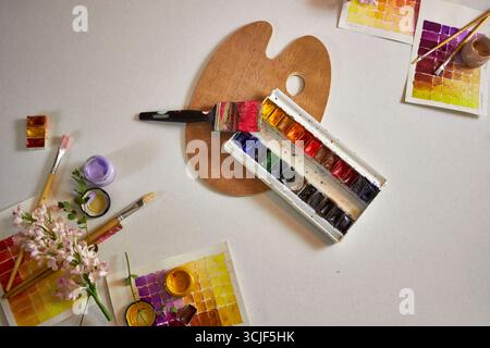 Künstlerische flache Lay mit Aquarellpalette, Pinseln, Farbmustern, Blumen und einer Holzpalette auf hellem Hintergrund. Kreativer Arbeitsbereich mit brillanter Leistung Stockfoto