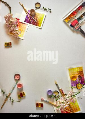 Flach mit Aquarellfarben, Pinseln, Farbmustern und frischen Blumen auf hellem Hintergrund. Künstlerischer Arbeitsbereich mit kreativen Werkzeugen und leerem SP Stockfoto