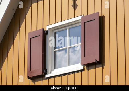 Offenes Fenster mit roten Fensterläden, die den blauen Himmel an einer orangen Holzwand eines Hauses reflektieren Stockfoto