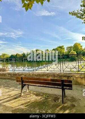 Holzbank mit Blick auf das Barrage des Forges in Audincourt, Frankreich, mit dekorativen Geländern und Reflexen von grünen Bäumen auf dem ruhigen Wasser Stockfoto
