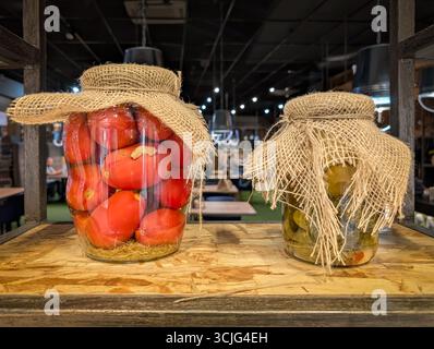 Zwei große Gläser mit traditionellen, hausgemachten eingelegten roten Tomaten und grünen Gurken stehen auf einem rustikalen Holzregal. Stockfoto