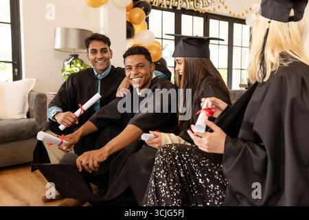 Absolventen in Mützen und Kleidern feiern Erfolge mit Diplomen zu Hause Stockfoto