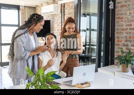 Im Büro feuern sich verschiedene Geschäftsfrauen um den Laptop und feiern gemeinsam Erfolge Stockfoto