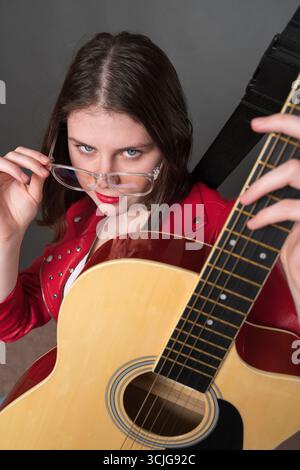 Die junge Gitarristin hält eine klassische Gitarre mit sechs Saiten und schaut in die Kamera, senkt ihre Brille und hält sie mit einer Hand am Tempel. Vertrauen Sie Sich An Stockfoto