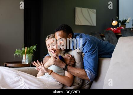 Afroamerikaner, der eine Frau auf der Couch überrascht, beide sehen fröhlich aus Stockfoto